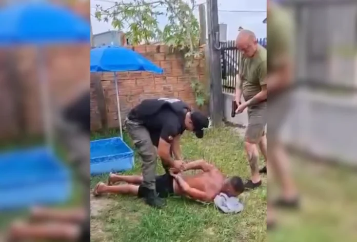 Foragido com pena de 52 anos é preso em Araranguá Foto de uma abordagem policial em um gramado residencial. Um policial civil, usando colete balístico e uniforme preto, algema um homem deitado de bruços no chão. Um segundo policial está em pé ao lado, dando cobertura à ação. Ao fundo, vê-se um guarda-sol azul e uma pequena piscina infantil. O detido é um foragido condenado a 52 anos de prisão.