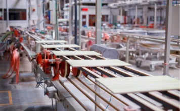 Força global: Sul de SC exporta US$ 160 mi; cerâmica é referência Linha de produção automatizada de uma fábrica de cerâmica. A imagem em perspectiva mostra ladrilhos e pisos claros sendo transportados rapidamente por uma esteira de roletes em tons de metal e laranja. O ambiente é industrial e amplo, com equipamentos e tubulações visíveis ao fundo.