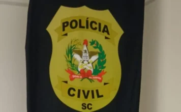 Gaivota: Foragido do RS é preso com vida ‘normal’ em SC Bandeira ou estandarte da Polícia Civil de Santa Catarina (PC/SC) em um fundo claro. O brasão central é um escudo amarelo com uma estrela branca no meio, rodeada por espigas de trigo e um galho de café, além das palavras "Estado de Santa Catarina" em vermelho. Acima do brasão, lê-se "POLÍCIA" e abaixo "CIVIL SC", tudo sobre um fundo de tecido escuro. A imagem representa a instituição responsável pela prisão do foragido do Rio Grande do Sul.