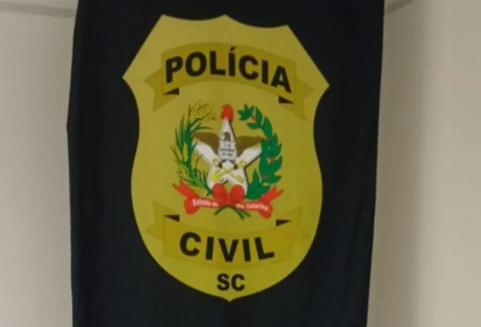 Gaivota Foragido do RS é preso com vida 'normal' em SC Bandeira ou estandarte da Polícia Civil de Santa Catarina (PC/SC) em um fundo claro. O brasão central é um escudo amarelo com uma estrela branca no meio, rodeada por espigas de trigo e um galho de café, além das palavras "Estado de Santa Catarina" em vermelho. Acima do brasão, lê-se "POLÍCIA" e abaixo "CIVIL SC", tudo sobre um fundo de tecido escuro. A imagem representa a instituição responsável pela prisão do foragido do Rio Grande do Sul.