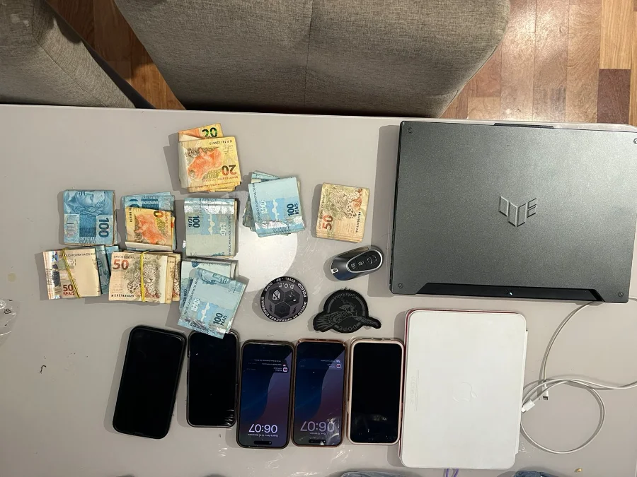 Golpistas são presos por crimes cibernéticos em quatro estados Visão superior de uma mesa onde estão dispostos objetos apreendidos em uma operação policial contra crimes cibernéticos. Os itens incluem várias cédulas de Real (notas de R$ 100, R$ 50 e R$ 20) espalhadas em pequenos montes, quatro smartphones com telas ligadas, um tablet branco, uma chave de carro e um notebook gamer preto fechado (com o logo