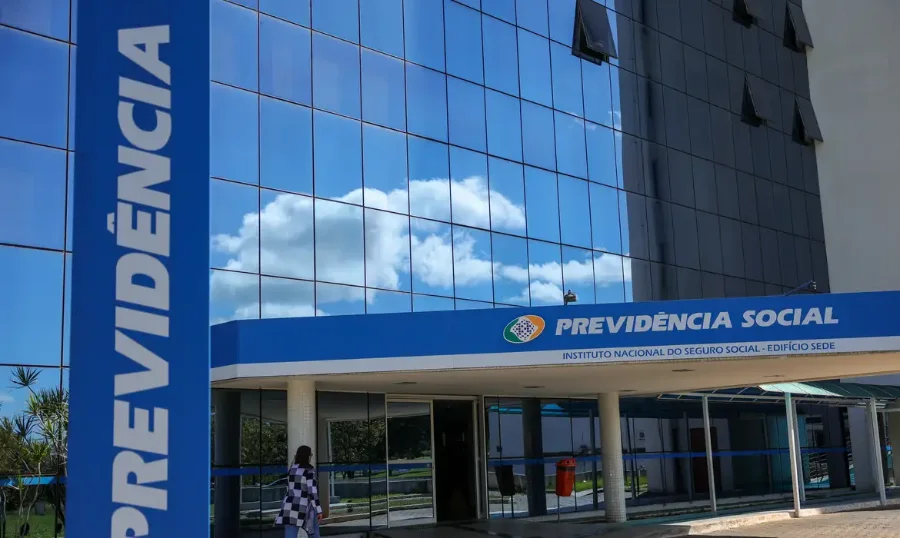 Fachada de um prédio da Previdência Social em um dia de sol. O edifício tem uma estrutura moderna, com uma parede envidraçada que reflete o céu azul com nuvens brancas. Na parte superior da entrada, há uma faixa azul com o logotipo e o nome 