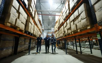 Imetro-SC lidera ranking nacional de fiscalização em 2025 Três fiscais do Imetro-SC, vestindo coletes marinho, caminham por um corredor de um grande armazém logístico com prateleiras altas repletas de caixas de papelão; ilustra a liderança nacional em fiscalização em 2025.