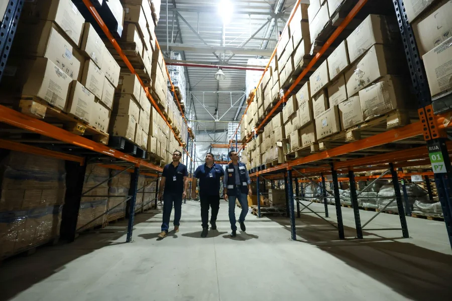 Três fiscais do Imetro-SC, vestindo coletes marinho, caminham por um corredor de um grande armazém logístico com prateleiras altas repletas de caixas de papelão; ilustra a liderança nacional em fiscalização em 2025.