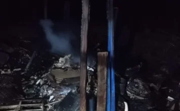 Incêndio destrói depósito de madeira em Balneário Gaivota Escombros carbonizados e fumaça no chão de um depósito de madeira destruído por incêndio durante a noite, com vigas azuis verticais aparentes.