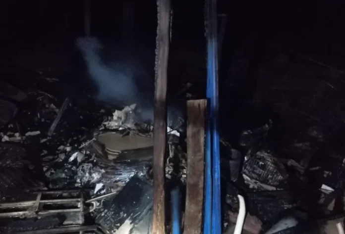 Incêndio destrói depósito de madeira em Balneário Gaivota Escombros carbonizados e fumaça no chão de um depósito de madeira destruído por incêndio durante a noite, com vigas azuis verticais aparentes.