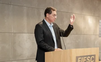 Indústria: China exige parcerias e IA; inércia é o maior risco Julio Damião, presidente do Ibef-MG, em pé em um púlpito de madeira com a logomarca da FIESC, gesticula com a mão direita enquanto profere uma palestra. Ele veste um terno preto e camisa azul clara. Ao fundo, uma parede revestida de painéis cinzas em uma sala de conferências da Federação das Indústrias.