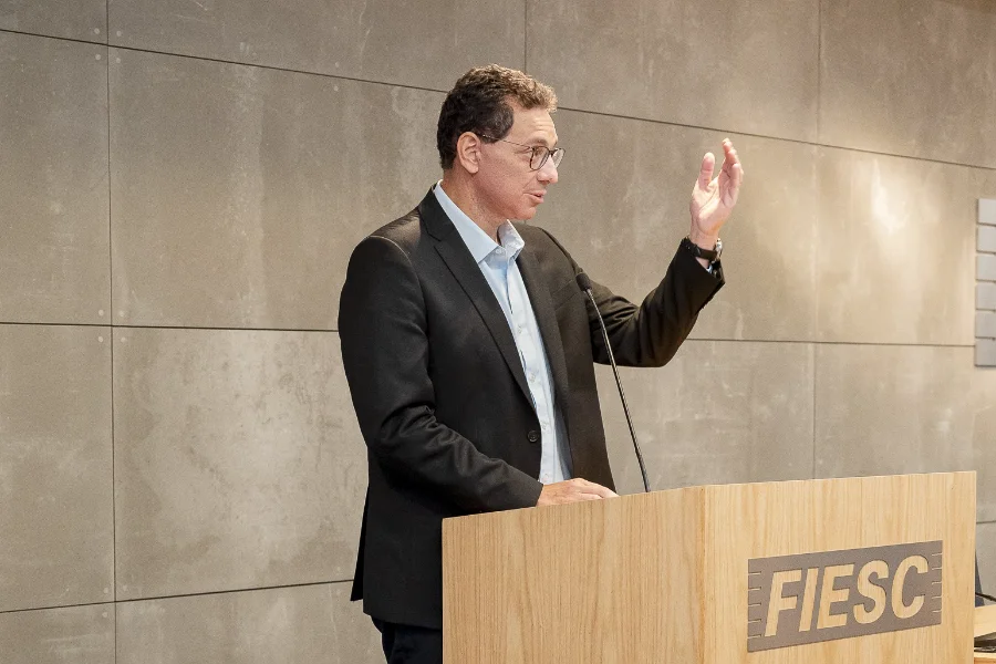 Indústria China exige parcerias e IA; inércia é o maior risco Julio Damião, presidente do Ibef-MG, em pé em um púlpito de madeira com a logomarca da FIESC, gesticula com a mão direita enquanto profere uma palestra. Ele veste um terno preto e camisa azul clara. Ao fundo, uma parede revestida de painéis cinzas em uma sala de conferências da Federação das Indústrias.