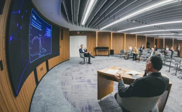 Indústria de SC cresce 2,8% e supera média nacional em 2025 Imagem de uma apresentação da FIESC em uma sala de reunião moderna e semicircular. Um homem de costas está sentado à mesa, gesticulando. À frente, uma tela curva exibe gráficos de crescimento. O público está sentado em cadeiras dispostas em semicírculo.