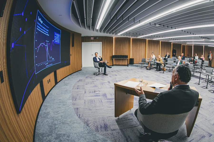 Imagem de uma apresentação da FIESC em uma sala de reunião moderna e semicircular. Um homem de costas está sentado à mesa, gesticulando. À frente, uma tela curva exibe gráficos de crescimento. O público está sentado em cadeiras dispostas em semicírculo.