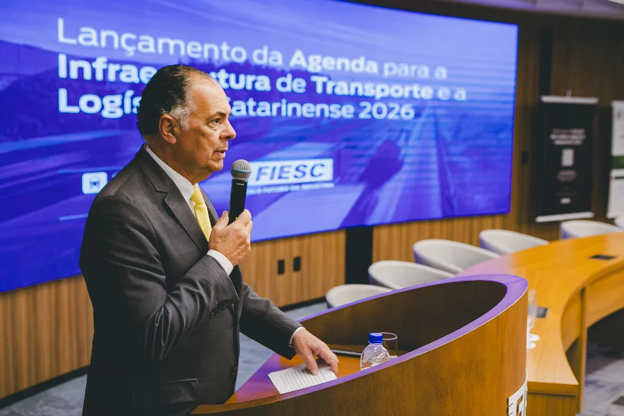 O presidente da FIESC, Gilberto Seleme, discursa no púlpito durante o lançamento da Agenda Estratégica para Infraestrutura, com o tema exibido ao fundo.