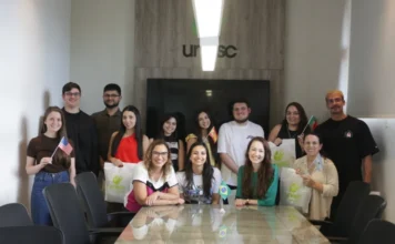 Internacionalização: Unesc envia alunos para Europa e Américas Grupo de estudantes da Unesc posa com bandeiras de países como EUA, Brasil e Bulgária em uma sala de reunião com o logo da universidade ao fundo.