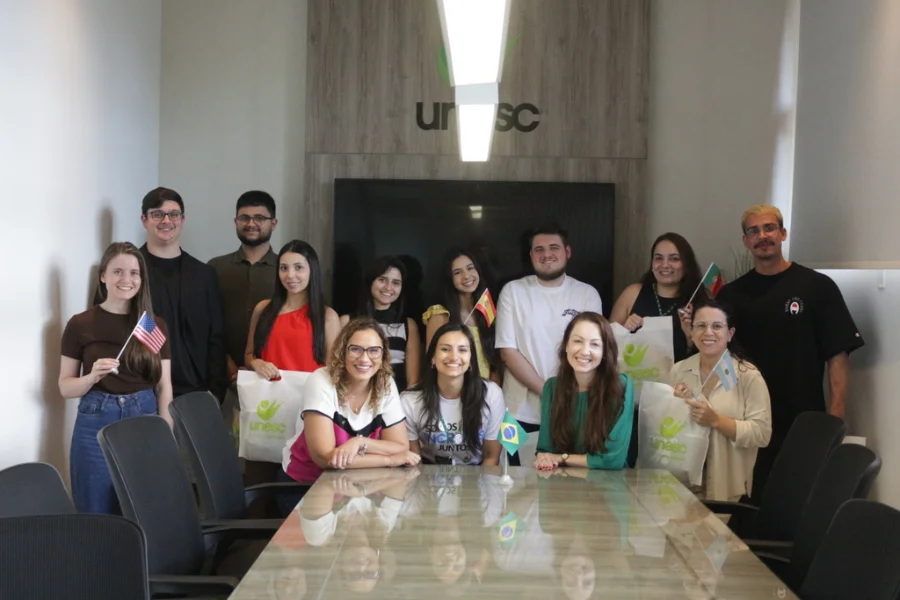 Grupo de estudantes da Unesc posa com bandeiras de países como EUA, Brasil e Bulgária em uma sala de reunião com o logo da universidade ao fundo.