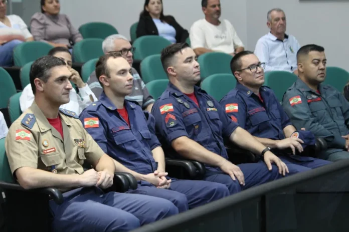Jiu-Jitsu e Bombeiro Mirim recebem homenagem em Forquilhinha Grupo de bombeiros militares sentados na primeira fila de um auditório durante sessão de homenagem na Câmara de Forquilhinha.