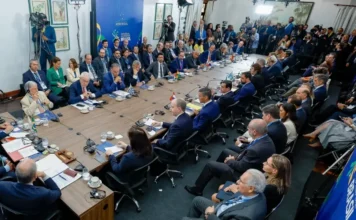 Lula alerta Mercosul sobre risco de conflito na Venezuela Fotografia em ângulo alto de uma grande sala de reuniões durante a Cúpula do Mercosul em Foz do Iguaçu. Chefes de Estado e delegados estão sentados ao redor de uma mesa em formato de "U". Ao fundo, um painel azul exibe a logomarca do Mercosul e o texto "Presidência Pro Tempore Brasil". O presidente Lula aparece à cabeceira da mesa.