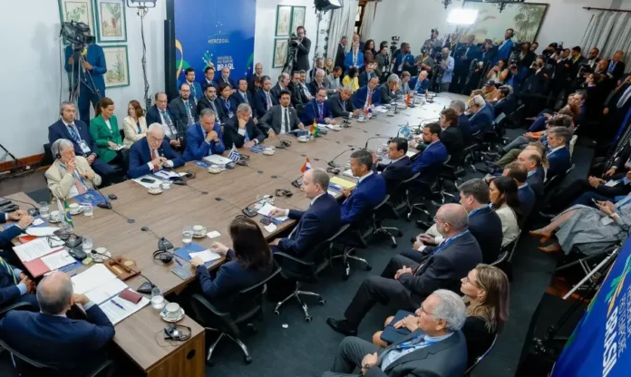 Lula alerta Mercosul sobre risco de conflito na Venezuela Fotografia em ângulo alto de uma grande sala de reuniões durante a Cúpula do Mercosul em Foz do Iguaçu. Chefes de Estado e delegados estão sentados ao redor de uma mesa em formato de "U". Ao fundo, um painel azul exibe a logomarca do Mercosul e o texto "Presidência Pro Tempore Brasil". O presidente Lula aparece à cabeceira da mesa.