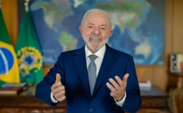 Lula celebra menor desigualdade com isenção do IR de R$ 5 mil Presidente Luiz Inácio Lula da Silva, de terno azul, sorrindo e gesticulando em um pronunciamento, com bandeiras do Brasil e mapa-múndi ao fundo.