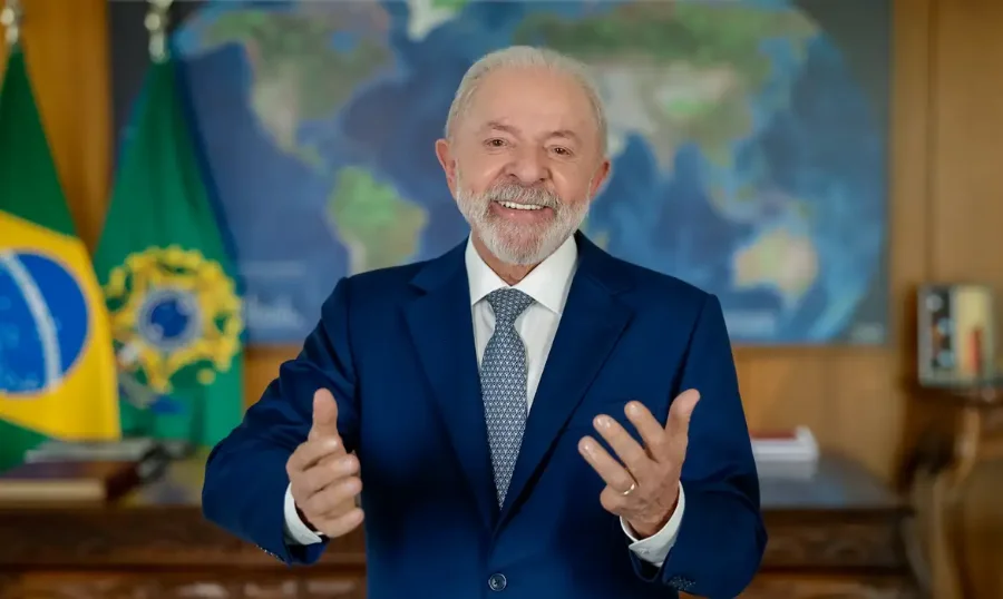 Presidente Luiz Inácio Lula da Silva, de terno azul, sorrindo e gesticulando em um pronunciamento, com bandeiras do Brasil e mapa-múndi ao fundo.