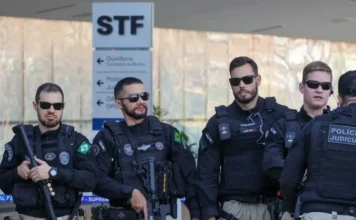 Lula sanciona lei que cria a carreira de Polícia Judicial Quatro agentes da Polícia Judicial em pé, vestindo uniformes pretos táticos com coletes balísticos e óculos escuros. Dois deles portam armamento longo. Ao fundo, uma placa branca indica as direções para o STF (Supremo Tribunal Federal) e a Ouvidoria. A imagem ilustra a sanção da lei que reorganiza a carreira e define novas nomenclaturas como Agentes e Inspetores.