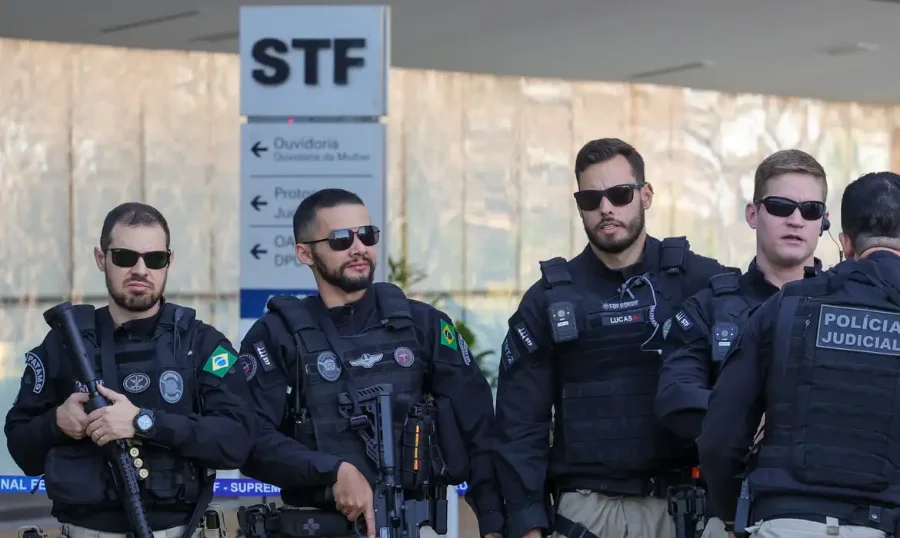 Quatro agentes da Polícia Judicial em pé, vestindo uniformes pretos táticos com coletes balísticos e óculos escuros. Dois deles portam armamento longo. Ao fundo, uma placa branca indica as direções para o STF (Supremo Tribunal Federal) e a Ouvidoria. A imagem ilustra a sanção da lei que reorganiza a carreira e define novas nomenclaturas como Agentes e Inspetores.