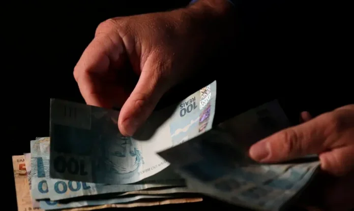 MP libera saque do FGTS retido para demitidos desde 2020 Fotografia em close-up das mãos de uma pessoa manuseando várias cédulas de 100 reais sobre um fundo escuro. A luz incide diretamente sobre o dinheiro, destacando a textura das notas. A imagem ilustra a liberação de R$ 7,8 bilhões do FGTS para trabalhadores demitidos desde 2020.