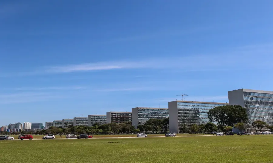 Vista panorâmica da Esplanada dos Ministérios, em Brasília, com uma fila de edifícios idênticos do lado direito e área verde em primeiro plano.