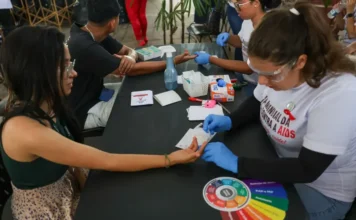 Ministro celebra redução de mortalidade por HIV no SUS Profissionais de saúde e voluntários em um estande aplicando testes rápidos de HIV no dedo de uma pessoa, durante campanha de conscientização.