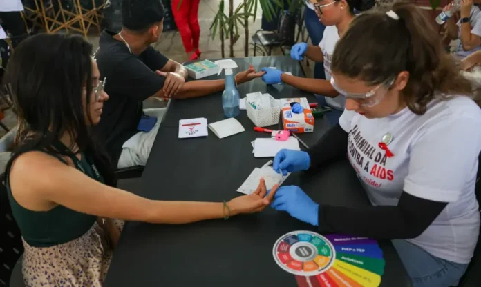 Ministro celebra redução de mortalidade por HIV no SUS Profissionais de saúde e voluntários em um estande aplicando testes rápidos de HIV no dedo de uma pessoa, durante campanha de conscientização.