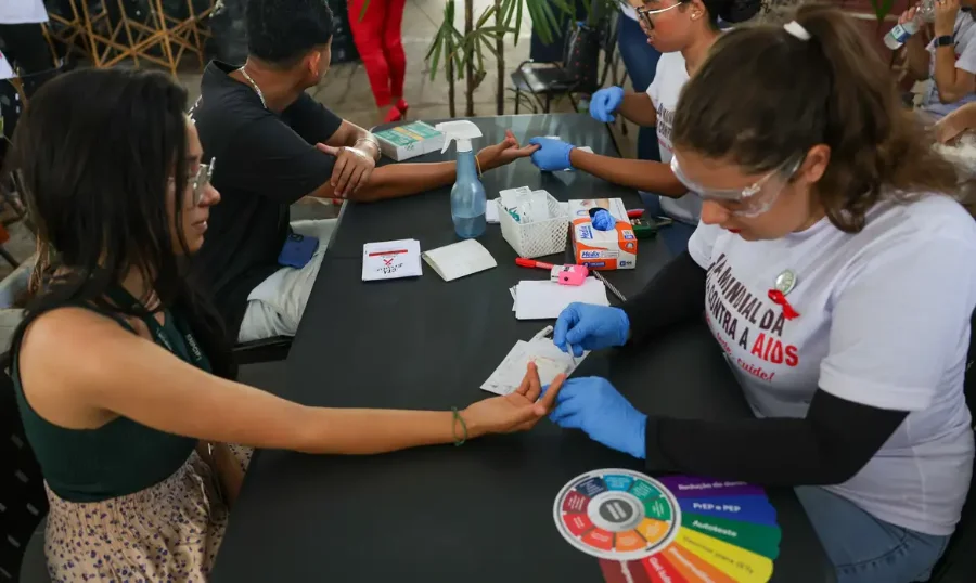 Ministro celebra redução de mortalidade por HIV no SUS Profissionais de saúde e voluntários em um estande aplicando testes rápidos de HIV no dedo de uma pessoa, durante campanha de conscientização.