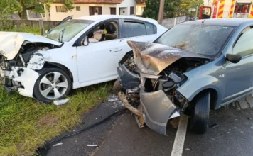 Morro da Fumaça: Colisão frontal é seguida de incêndio em carro Dois carros com as frentes completamente destruídas após colisão em rodovia, um branco e um cinza, com bombeiros ao fundo.