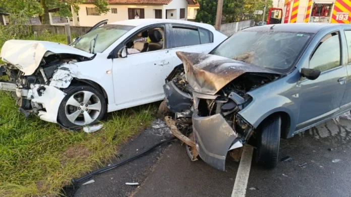 Morro da Fumaça Colisão frontal é seguida de incêndio em carro Dois carros com as frentes completamente destruídas após colisão em rodovia, um branco e um cinza, com bombeiros ao fundo.