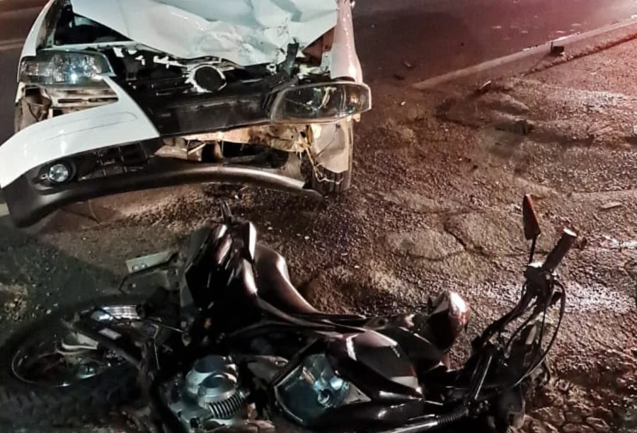Vistas noturnas da frente danificada de um carro branco (Volkswagen Gol) e de uma motocicleta preta deitada no asfalto após colisão frontal.