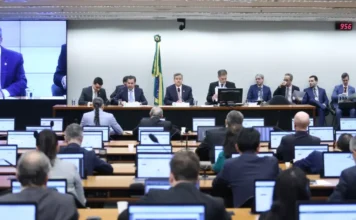 Orçamento 2026: CMO aprova salário mínimo de R$ 1.621 Mesa diretora da Comissão Mista de Orçamento (CMO) no Congresso Nacional. Parlamentares estão sentados à mesa sob uma bandeira do Brasil, enquanto monitores individuais à frente mostram outros membros da comissão. A reunião aprovou o salário mínimo de R$ 1.621 para 2026.