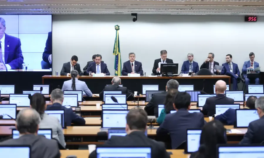 Mesa diretora da Comissão Mista de Orçamento (CMO) no Congresso Nacional. Parlamentares estão sentados à mesa sob uma bandeira do Brasil, enquanto monitores individuais à frente mostram outros membros da comissão. A reunião aprovou o salário mínimo de R$ 1.621 para 2026.