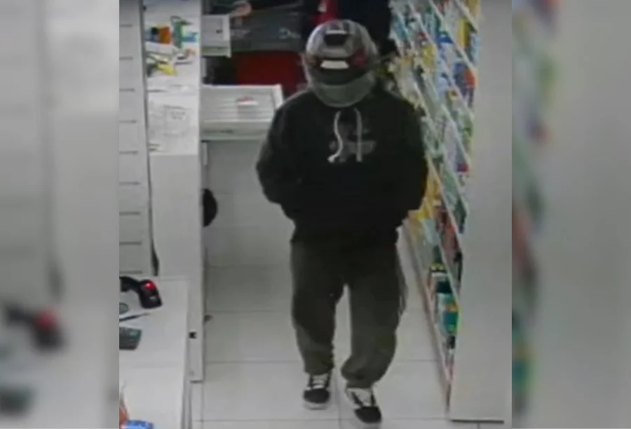 Polícia Civil indicia homem por cinco assaltos em Criciúma Imagem de câmera de monitoramento mostrando suspeito usando capacete e blusa escura dentro de uma farmácia.