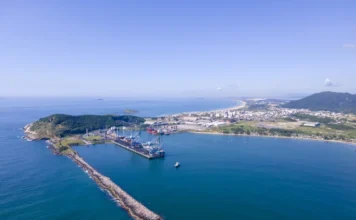 Porto de Imbituba recebe R$ 90,5 mi para reforço do molhe Foto aérea do Porto de Imbituba destacando o molhe de abrigo e guindastes de carga. Governo autoriza R$ 90,5 milhões para obras de recuperação e reforço estrutural, parte de um pacote de investimentos de R$ 300 milhões.