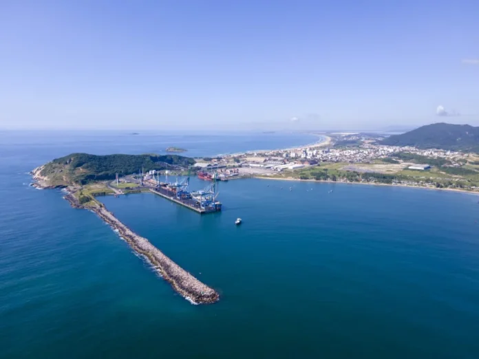 Porto de Imbituba recebe R$ 90,5 mi para reforço do molhe Foto aérea do Porto de Imbituba destacando o molhe de abrigo e guindastes de carga. Governo autoriza R$ 90,5 milhões para obras de recuperação e reforço estrutural, parte de um pacote de investimentos de R$ 300 milhões.