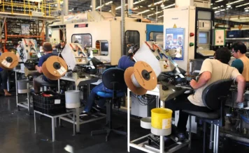 Produção industrial cresce 0,1% em outubro, puxada por petróleo Trabalhadores em uma linha de montagem industrial operando máquinas, com bobinas de fio em primeiro plano, representando a produção industrial.