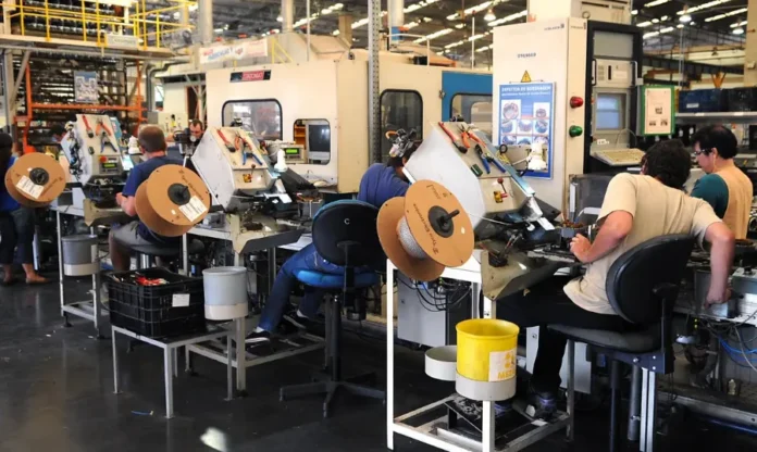 Produção industrial cresce 0,1% em outubro, puxada por petróleo Trabalhadores em uma linha de montagem industrial operando máquinas, com bobinas de fio em primeiro plano, representando a produção industrial.