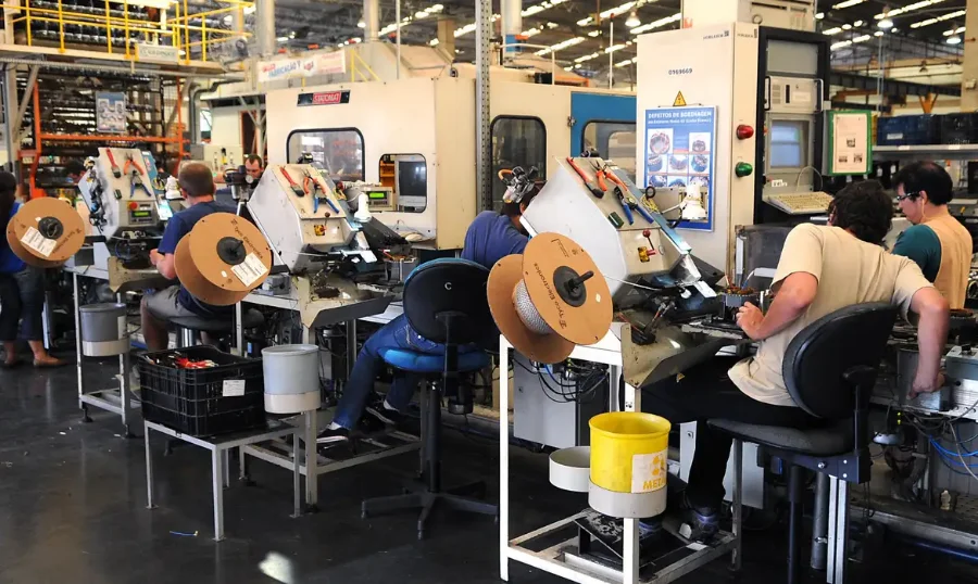 Trabalhadores em uma linha de montagem industrial operando máquinas, com bobinas de fio em primeiro plano, representando a produção industrial.