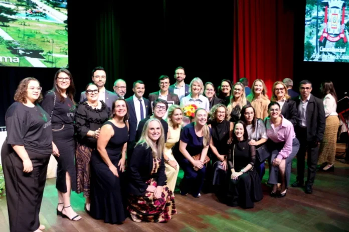 Reitora da Unesc recebe Cidadania Benemérita de Criciúma Gisele Silveira Coelho Lopes (reitora) e grupo de líderes e colegas posam juntos no palco do Teatro Elias Angeloni, celebrando a Cidadania Benemérita.