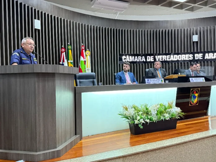 Rio Araranguá Vereadores debatem segurança e regulamentação Capitão de Corveta da Marinha, uniformizado, discursa no púlpito da Câmara de Vereadores de Araranguá durante sessão, com a mesa diretora e bandeiras ao fundo.