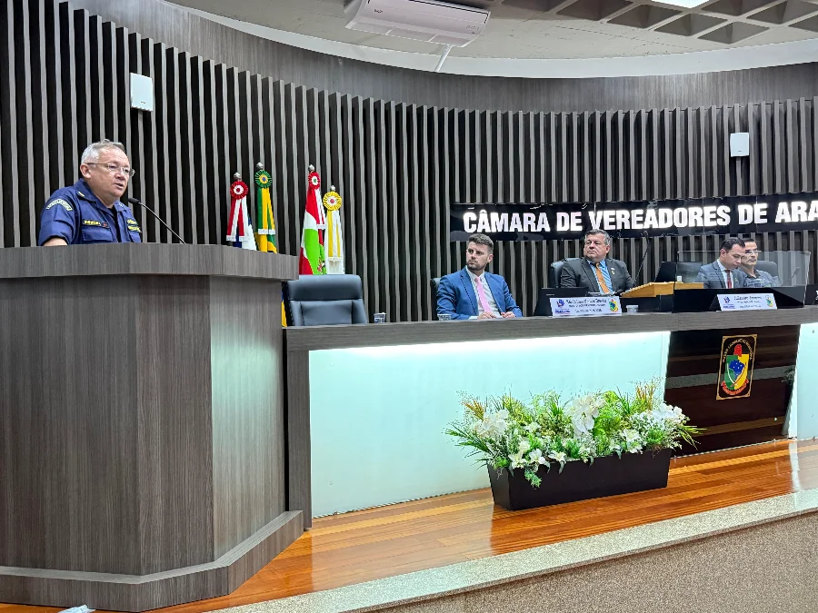 Rio Araranguá Vereadores debatem segurança e regulamentação Capitão de Corveta da Marinha, uniformizado, discursa no púlpito da Câmara de Vereadores de Araranguá durante sessão, com a mesa diretora e bandeiras ao fundo.