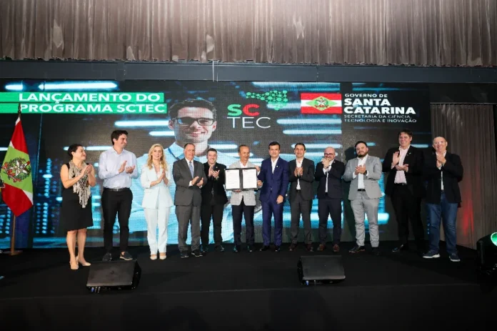 SC lança SCTEC com 155 mil vagas gratuitas em IA e tecnologia Lançamento do programa SCTEC do Governo de Santa Catarina. A imagem mostra uma cerimônia em palco, onde cerca de 10 homens de terno e uma mulher de vestido preto estão aplaudindo. Ao centro, dois homens trocam apertos de mão ou seguram um documento assinado. Atrás deles, um painel grande exibe o logo do programa "SCTEC" e o brasão do "Governo de Santa Catarina - Secretaria da Ciência, Tecnologia e Inovação", com a legenda "LANÇAMENTO DO PROGRAMA SCTEC" à esquerda. A cena representa a formalização do programa de 155 mil vagas gratuitas em tecnologia e IA.