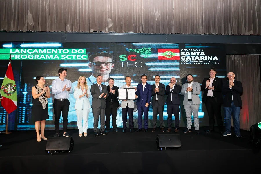 SC lança SCTEC com 155 mil vagas gratuitas em IA e tecnologia Lançamento do programa SCTEC do Governo de Santa Catarina. A imagem mostra uma cerimônia em palco, onde cerca de 10 homens de terno e uma mulher de vestido preto estão aplaudindo. Ao centro, dois homens trocam apertos de mão ou seguram um documento assinado. Atrás deles, um painel grande exibe o logo do programa
