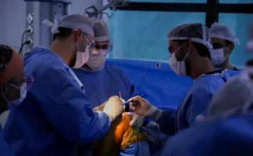 SC lidera cirurgias eletivas no país com marca histórica Equipe médica composta por três cirurgiões e um instrumentador, usando aventais azuis, máscaras e toucas descartáveis, realiza um procedimento cirúrgico em um joelho sob luzes de centro cirúrgico. A imagem ilustra a marca histórica de 1,2 milhão de procedimentos realizados em SC nos últimos três anos.
