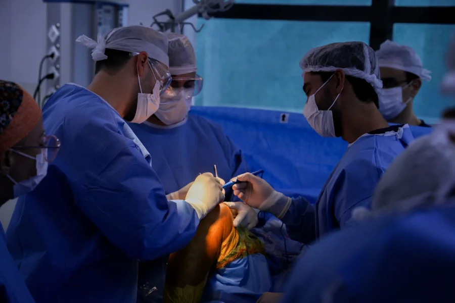Equipe médica composta por três cirurgiões e um instrumentador, usando aventais azuis, máscaras e toucas descartáveis, realiza um procedimento cirúrgico em um joelho sob luzes de centro cirúrgico. A imagem ilustra a marca histórica de 1,2 milhão de procedimentos realizados em SC nos últimos três anos.