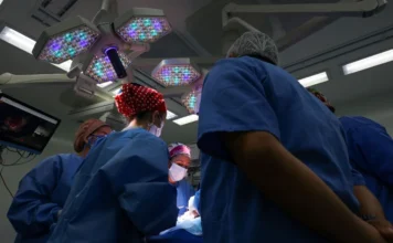 SC registra recorde histórico de 1,2 milhão de cirurgias Equipe médica em trajes cirúrgicos azuis, realizando um procedimento em sala de cirurgia, sob forte iluminação das luzes do teto.