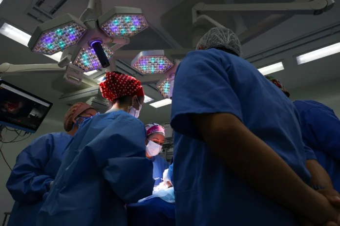 SC registra recorde histórico de 1,2 milhão de cirurgias Equipe médica em trajes cirúrgicos azuis, realizando um procedimento em sala de cirurgia, sob forte iluminação das luzes do teto.