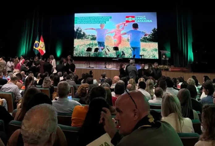 Santa Rosa do Sul garante R$ 676 mil para projeto de idosos Plenária lotada em evento com autoridades no palco, onde uma tela de LED exibe a mensagem "Projetos contemplados no Fundo da Infância e da Adolescência (FIA)".